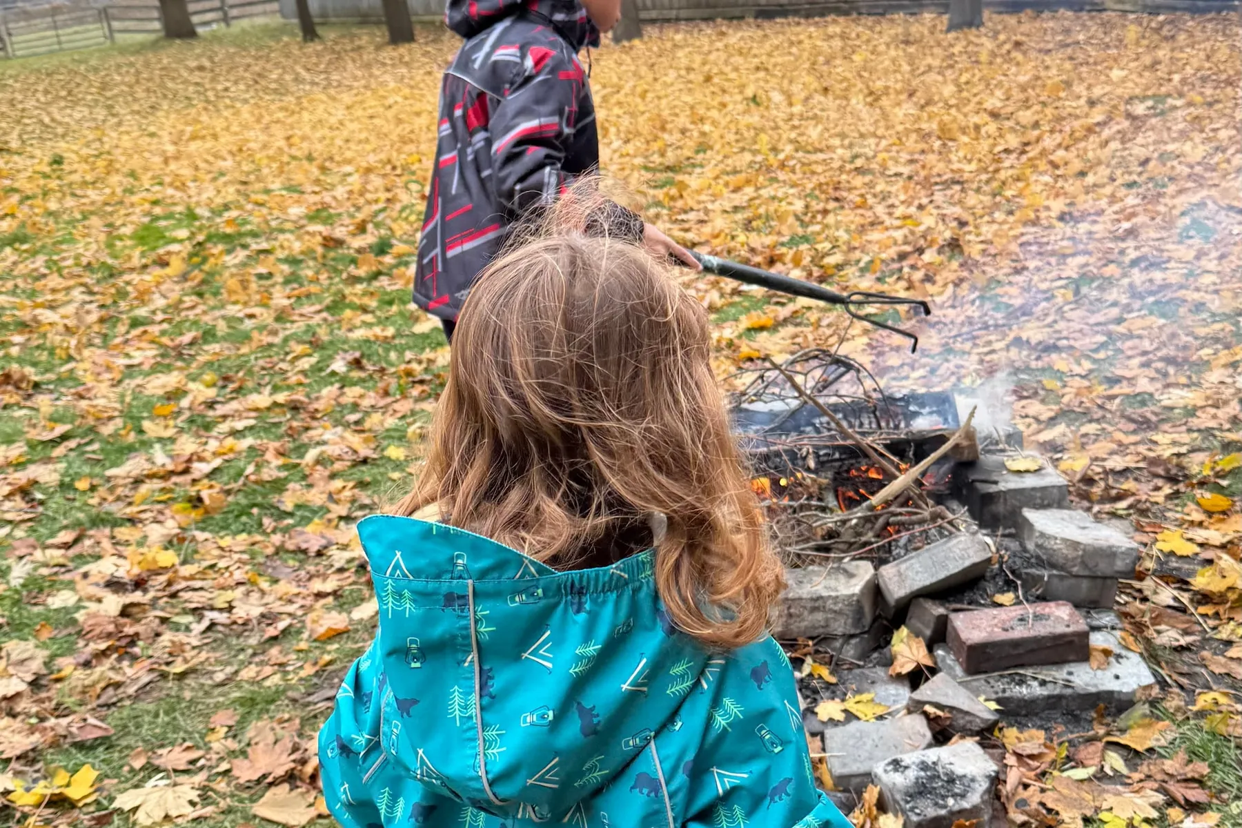 Two children in bright waterproofs beside a small fire ringed with stones, golden leaves covering the ground.