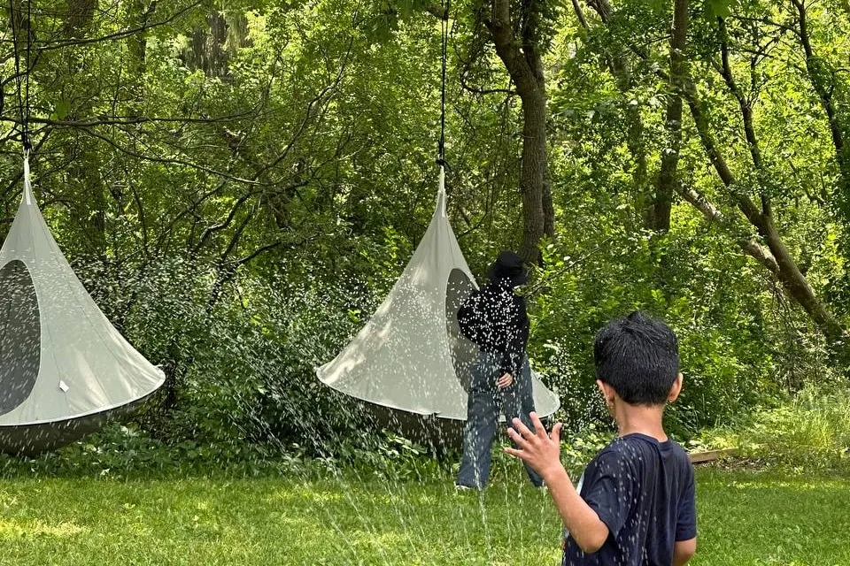 Two children playing in a sprinkler on the lawn beneath canvas hanging seats slung from the trees.