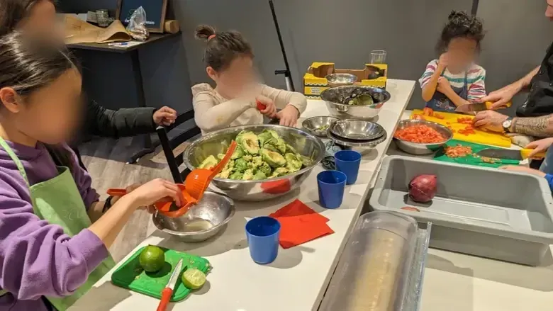 Children in aprons at a long table, peeling limes and chopping vegetables in the indoor kitchen.